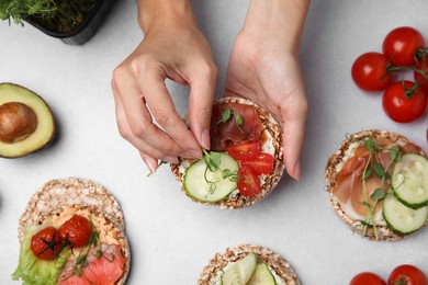 Photo of Woman adding microgreens onto crunchy buckwheat cakes with prosciutto, pieces of tomato and cucumber slice at white table, closeup