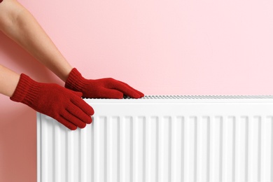 Woman in gloves warming hands on heating radiator near color wall. Space for text