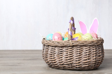 Photo of Wicker basket with Easter eggs and funny bunny ears on wooden table against light background, space for text