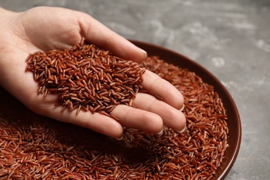 Woman holding grains near plate with brown rice on table, closeup