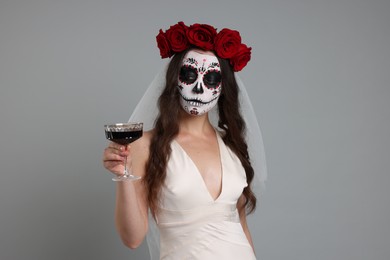 Young woman in scary bride costume with sugar skull makeup, flower crown and glass of wine on light grey background. Halloween celebration