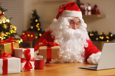 Photo of Santa Claus with Christmas gift using laptop at table in room