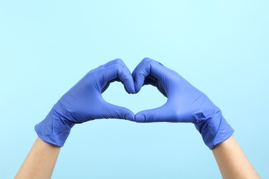 Person in medical gloves showing heart gesture on light blue background, closeup of hands