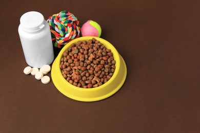 Photo of Bowl with dry pet food, bottle of vitamins and toys on brown background. Space for text