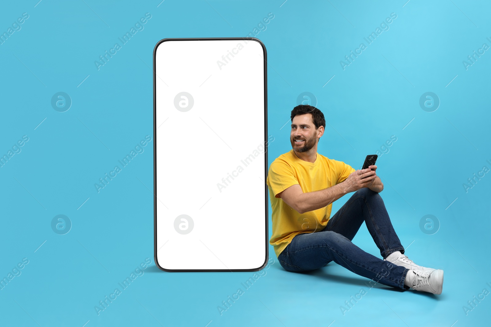 Image of Man with mobile phone sitting near huge device with empty screen on light blue background. Mockup for design