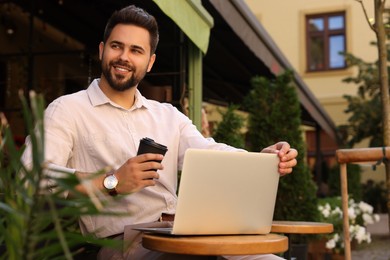 Photo of Handsome young man with cup of coffee working on laptop at table in outdoor cafe