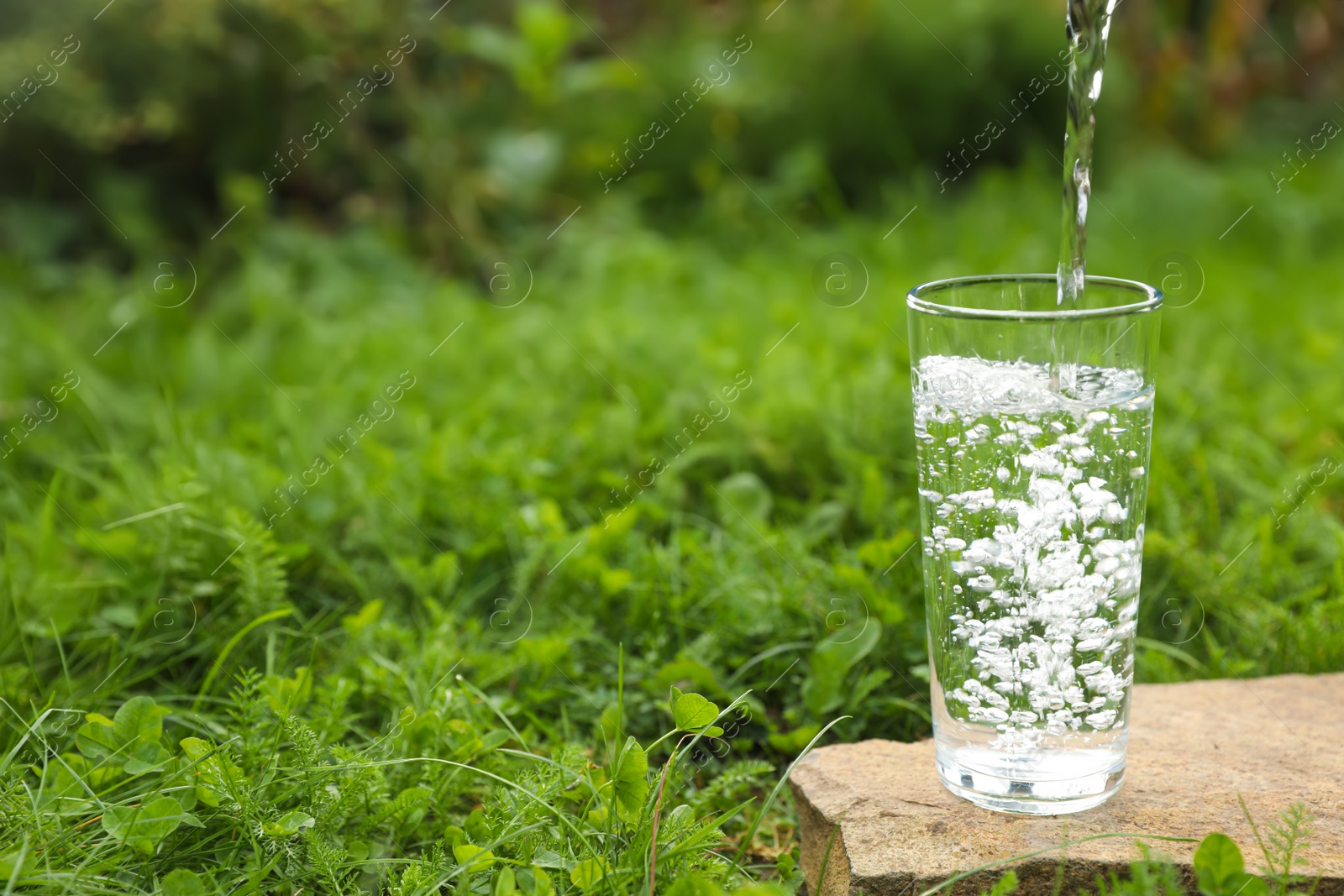 Photo of Pouring fresh water into glass on stone in green grass outdoors. Space for text