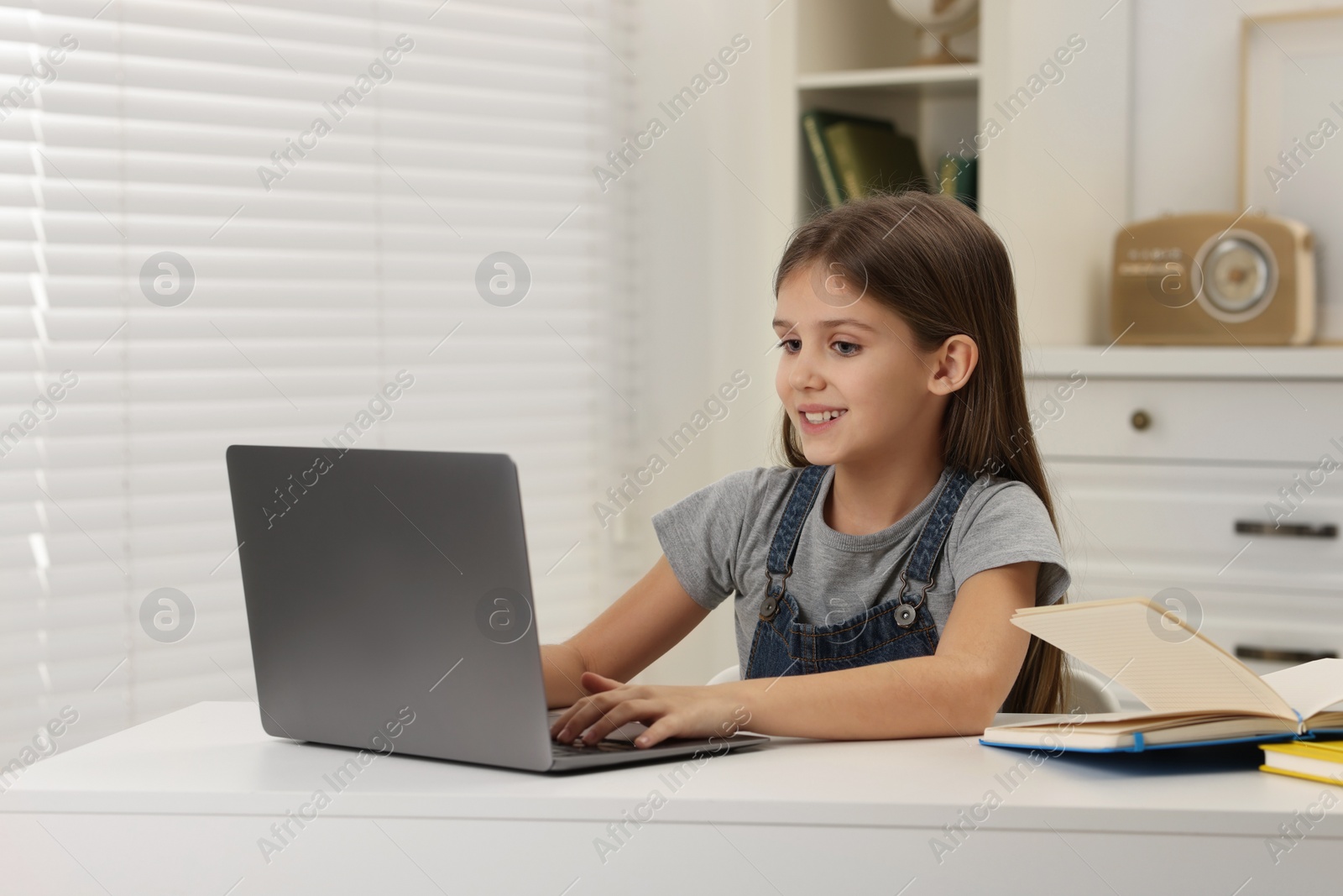 Photo of Cute girl using laptop at white table indoors