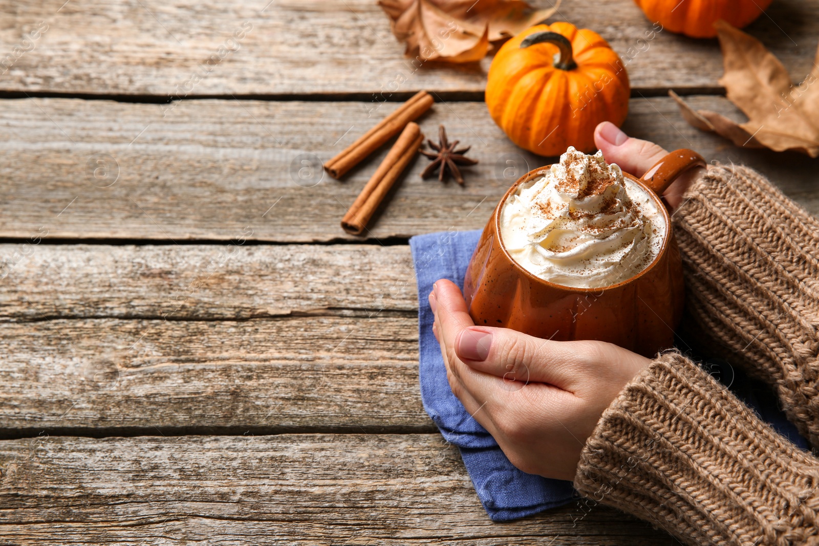 Photo of Woman holding mug of pumpkin spice latte with whipped cream at wooden table, closeup. Space for text