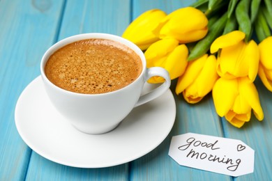 Photo of Cup of aromatic coffee, beautiful yellow tulips and Good Morning note on light blue wooden table