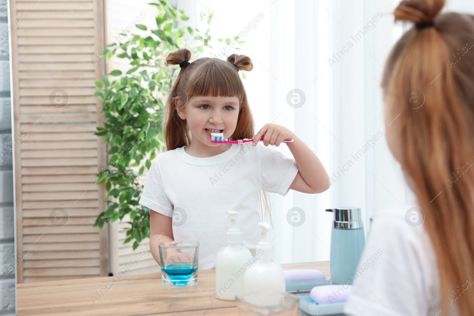 Photo of Little girl brushing teeth near mirror in bathroom at home