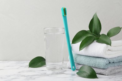 Photo of Plastic toothbrush, glass of water, towels and green leaves on white marble table, space for text