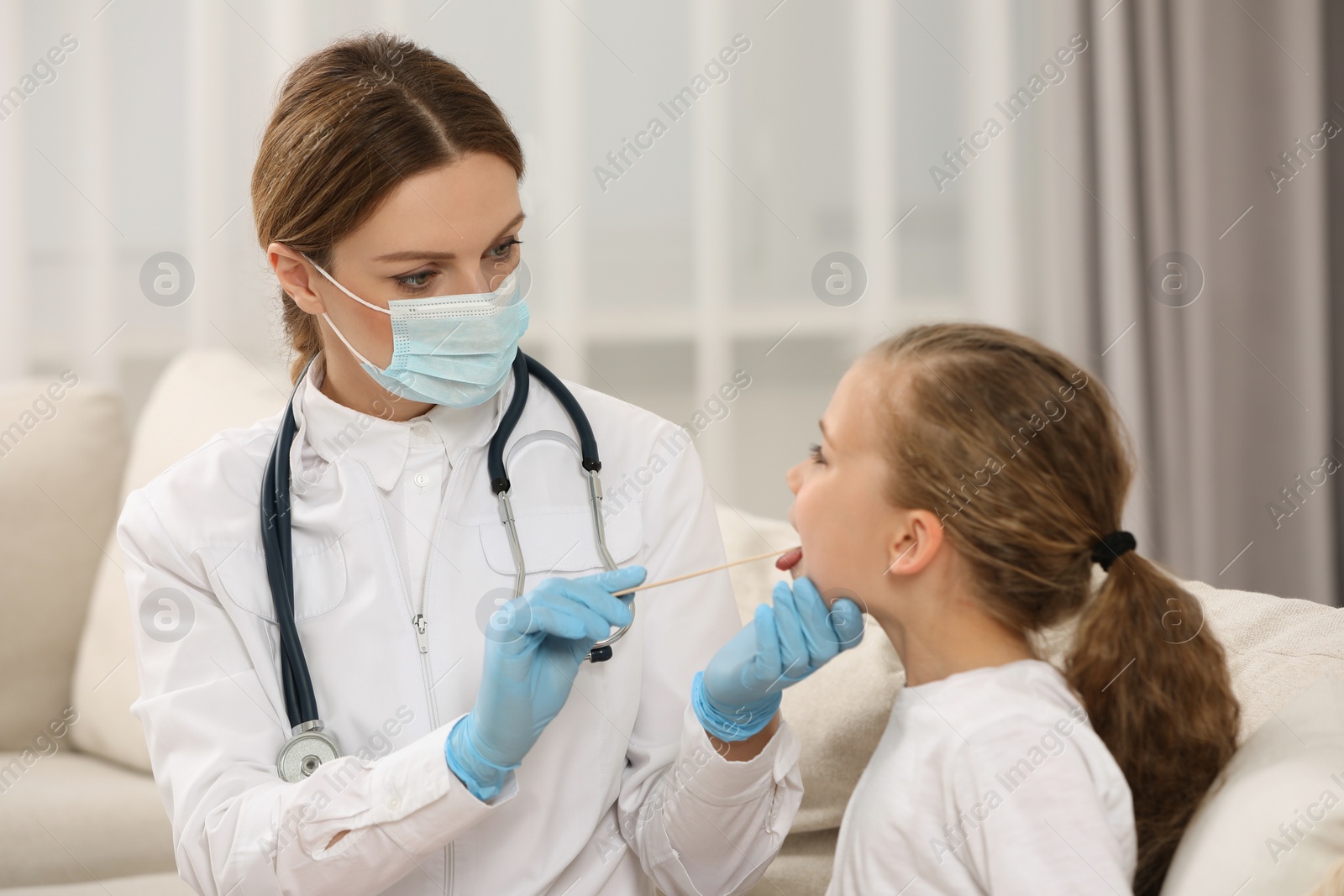 Photo of Doctor in medical mask examining girl`s oral cavity with tongue depressor indoors