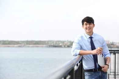 Photo of Young man with cup of coffee and laptop standing at pier. Joy in moment