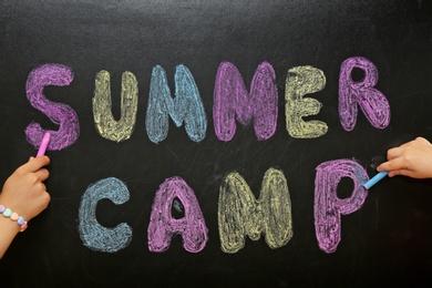 Photo of Children writing SUMMER CAMP with colorful chalk on blackboard, closeup