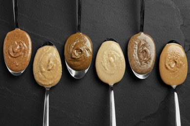 Tasty nut butters in spoons on dark gray textured table, flat lay