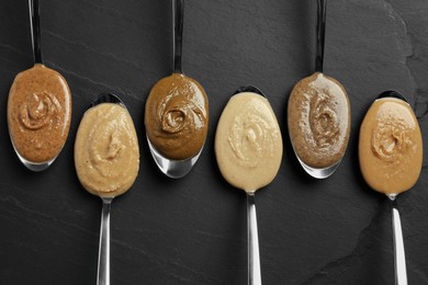 Photo of Tasty nut butters in spoons on dark gray textured table, flat lay