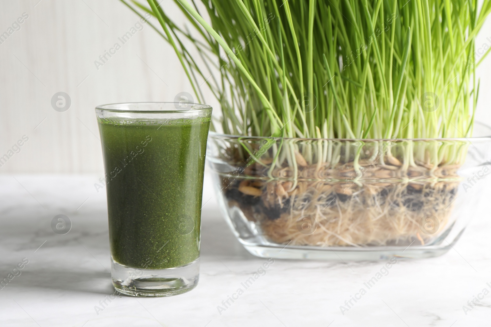 Photo of Wheat grass drink in shot glass and fresh sprouts on white marble table, closeup