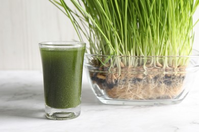 Photo of Wheat grass drink in shot glass and fresh sprouts on white marble table, closeup