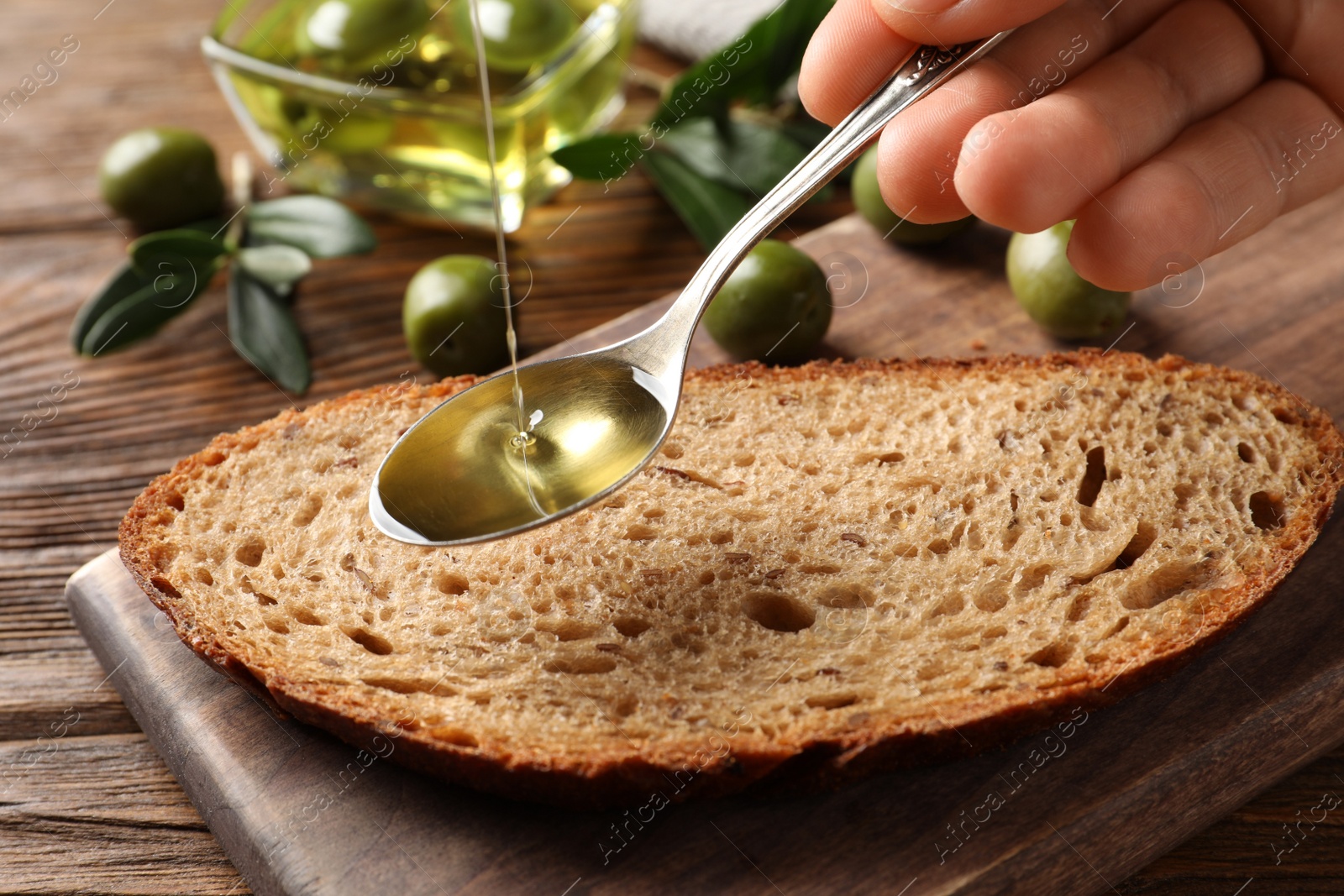 Photo of Woman pouring olive oil into spoon over bread at wooden table, closeup