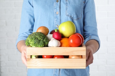 Photo of Woman holding wooden crate with fruits and vegetables at white brick wall, closeup