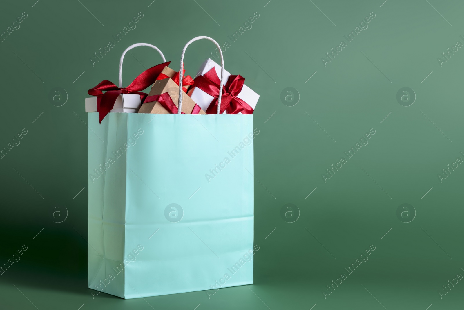 Photo of Turquoise paper shopping bag full of gift boxes on green background. Space for text