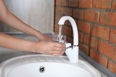 Woman filling glass with water from faucet in kitchen, closeup