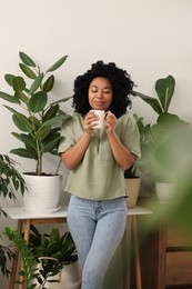 Photo of Relaxing atmosphere. Woman with cup of hot drink near beautiful houseplants in room
