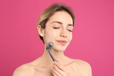 Young woman using metal face roller on pink background