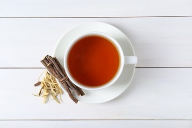 Aromatic licorice tea in cup and dried sticks of licorice root on white wooden table, top view