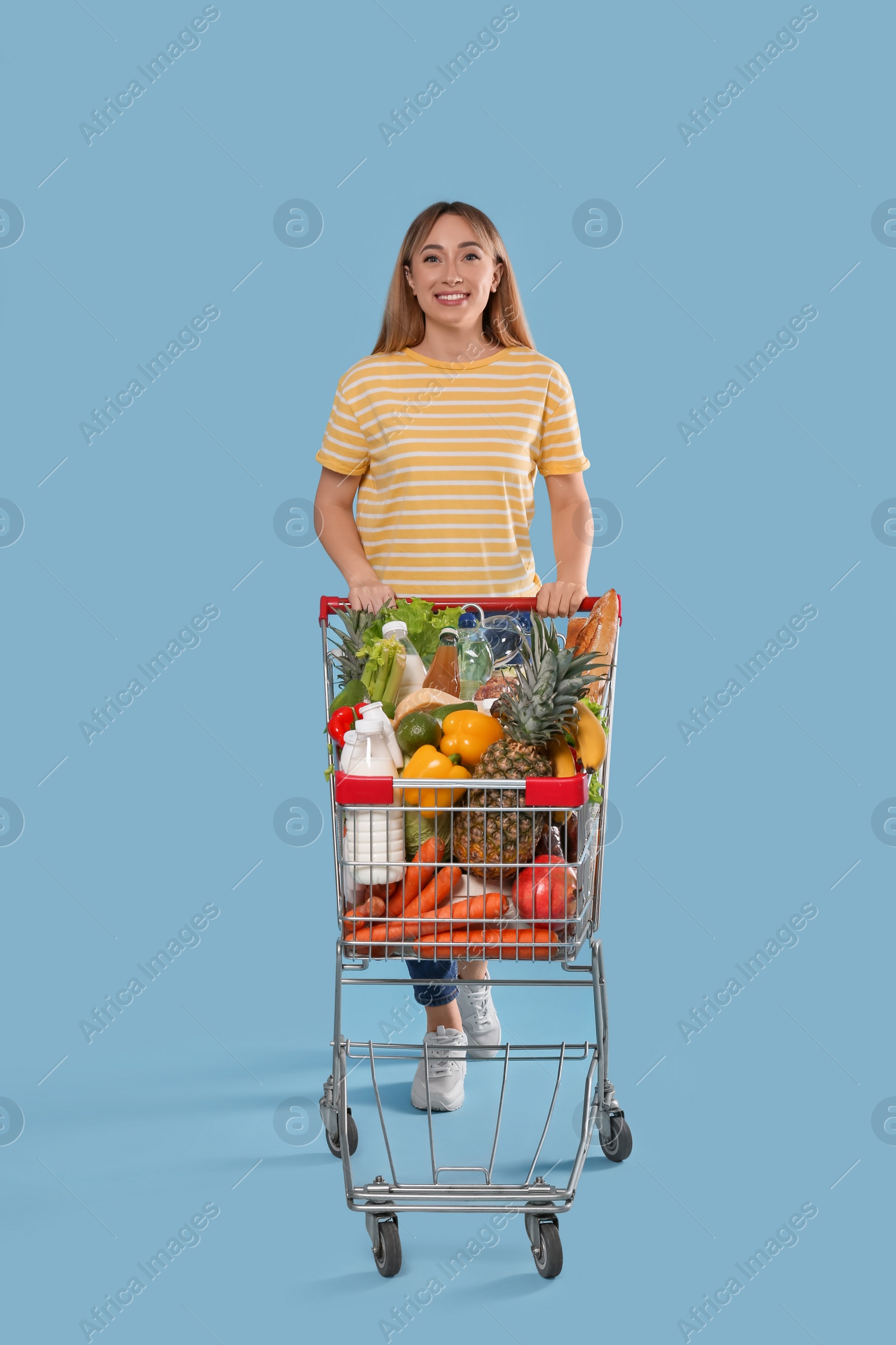 Photo of Young woman with shopping cart full of groceries on light blue background