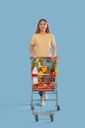 Photo of Young woman with shopping cart full of groceries on light blue background