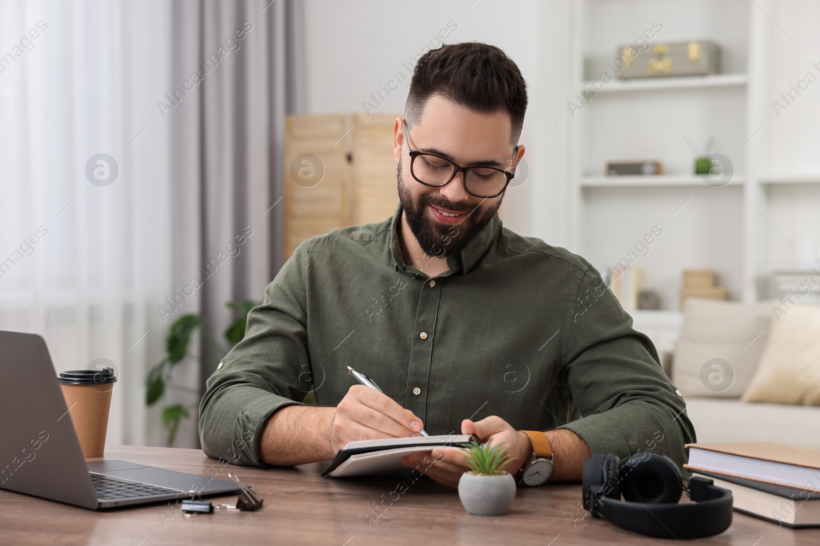 Photo of E-learning. Young man taking notes during online lesson at wooden table indoors