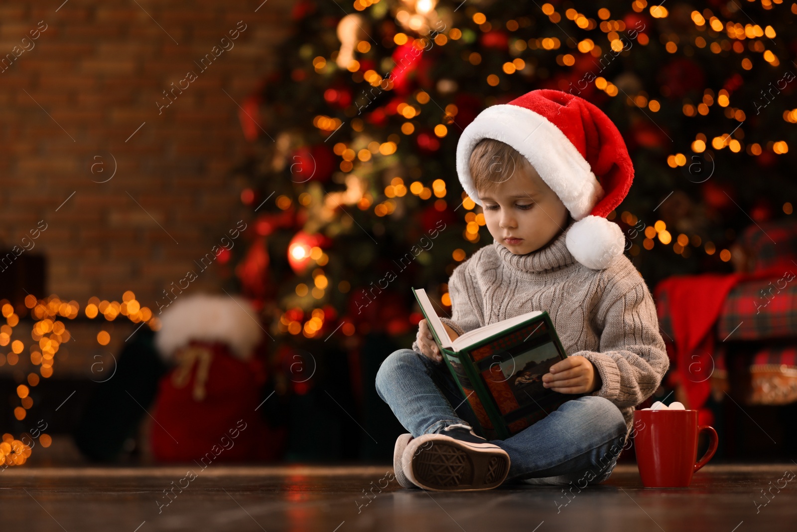 Photo of Little boy in Santa Claus cap reading book near Christmas tree at home