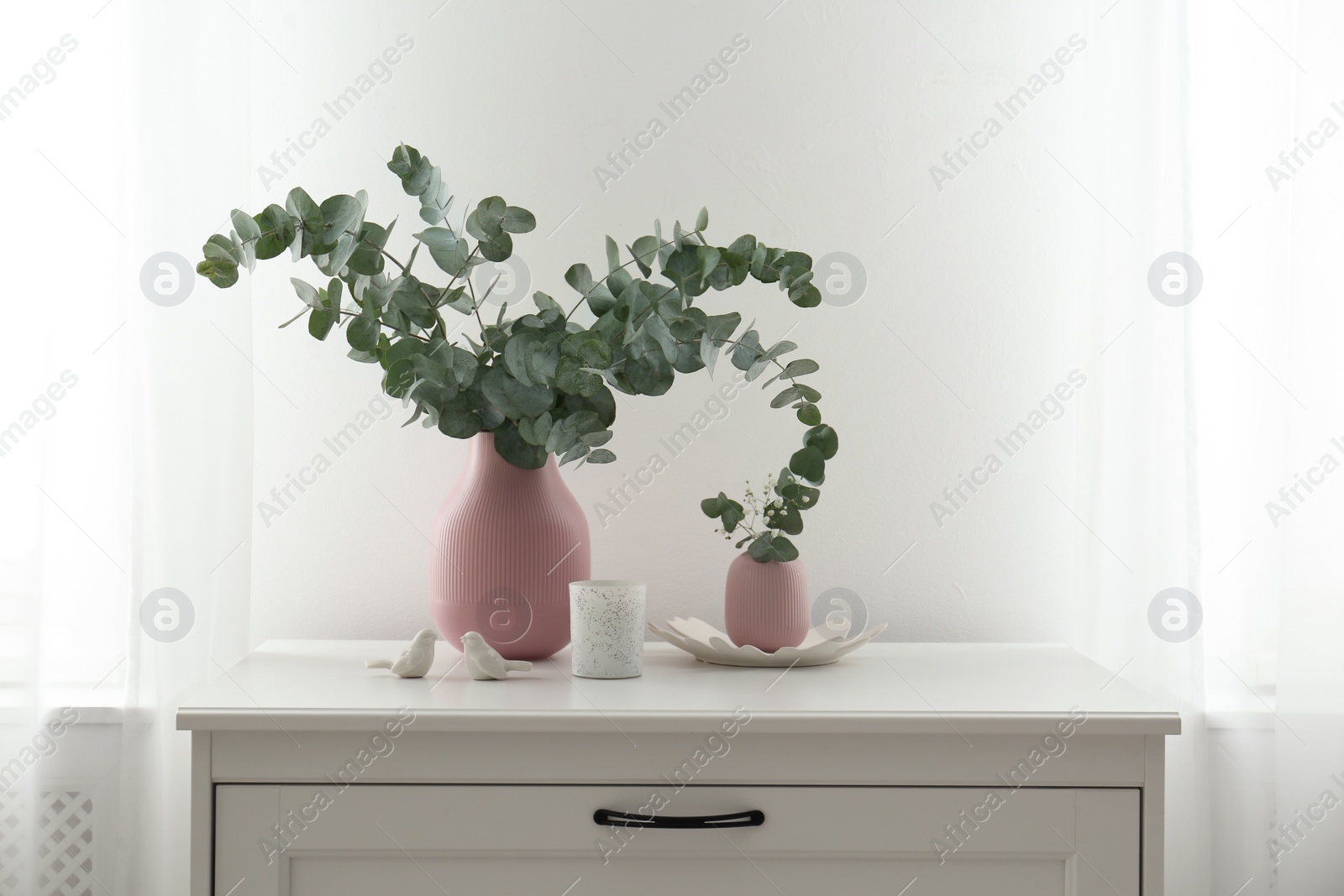 Photo of Beautiful eucalyptus branches, candle and bird figures on chest of drawers indoors. Interior element