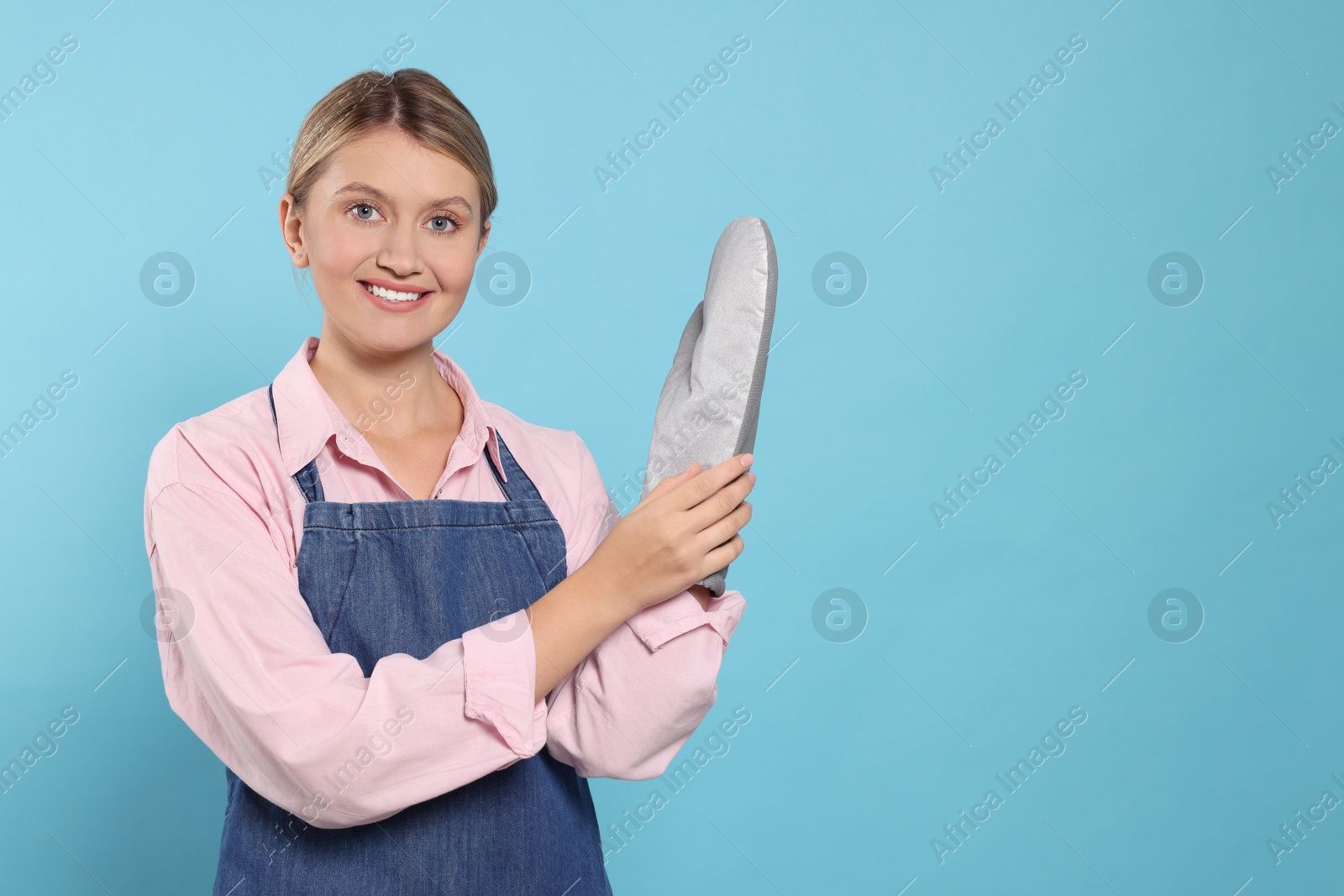 Photo of Beautiful young woman in denim apron and oven glove on light blue background. Space for text
