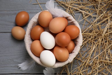 Photo of Fresh chicken eggs in bowl, feathers and dried straw on grey wooden table, flat lay