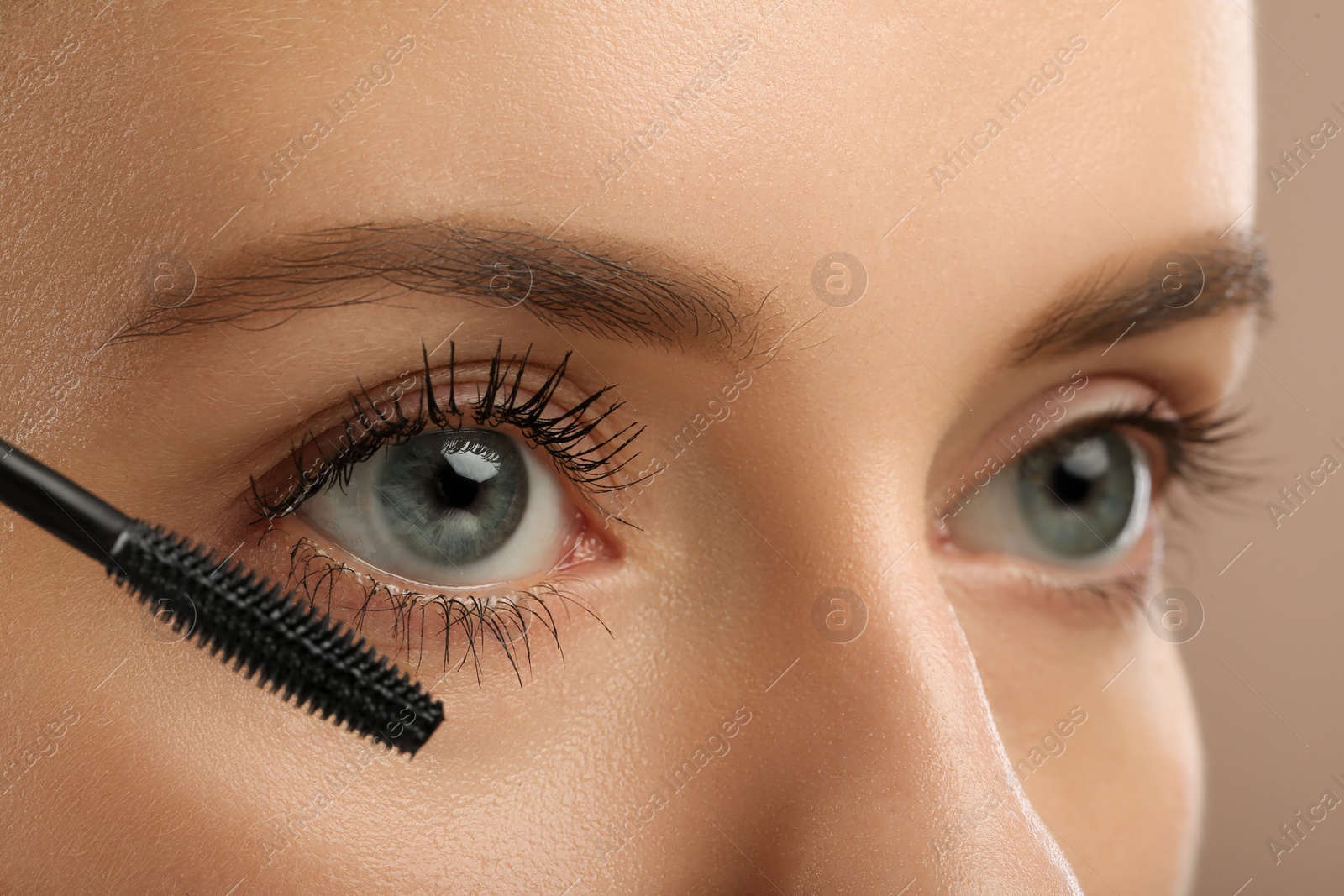 Photo of Woman applying mascara onto eyelashes against light brown background, closeup