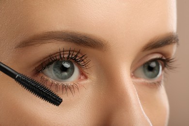 Woman applying mascara onto eyelashes against light brown background, closeup