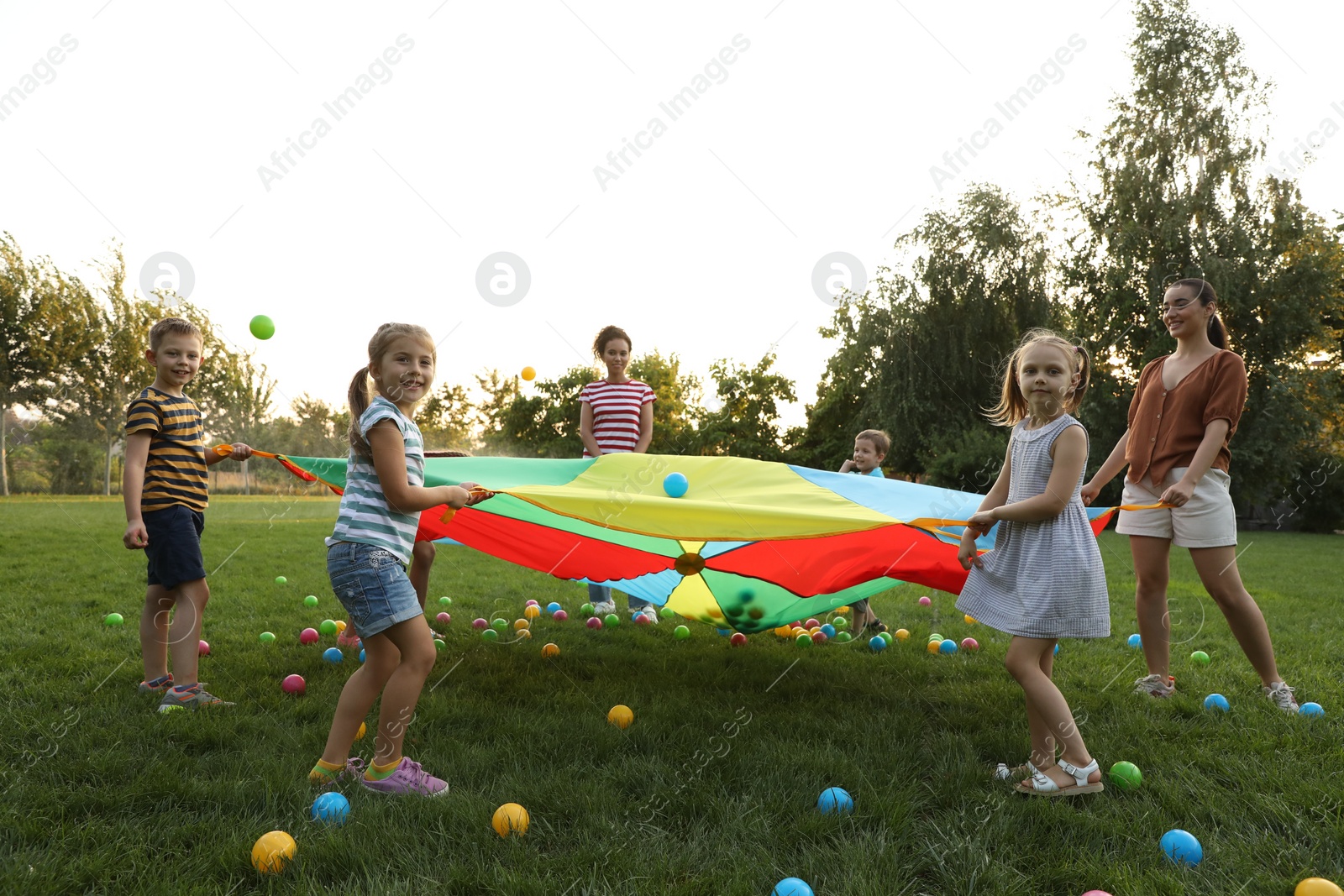 Photo of Group of children and teachers playing with rainbow playground parachute on green grass. Summer camp activity