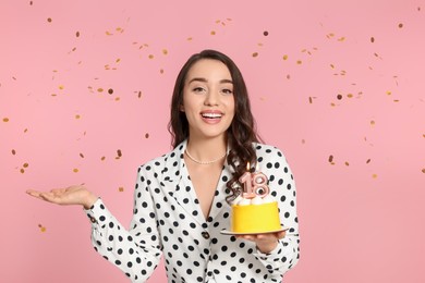 Coming of age party - 18th birthday. Smiling woman holding delicious cake with number shaped candles and catching confetti on pink background
