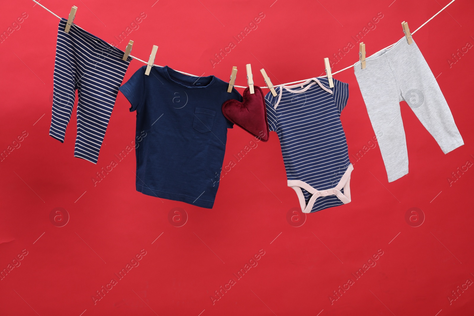 Photo of Different baby clothes and heart shaped cushion drying on laundry line against red background