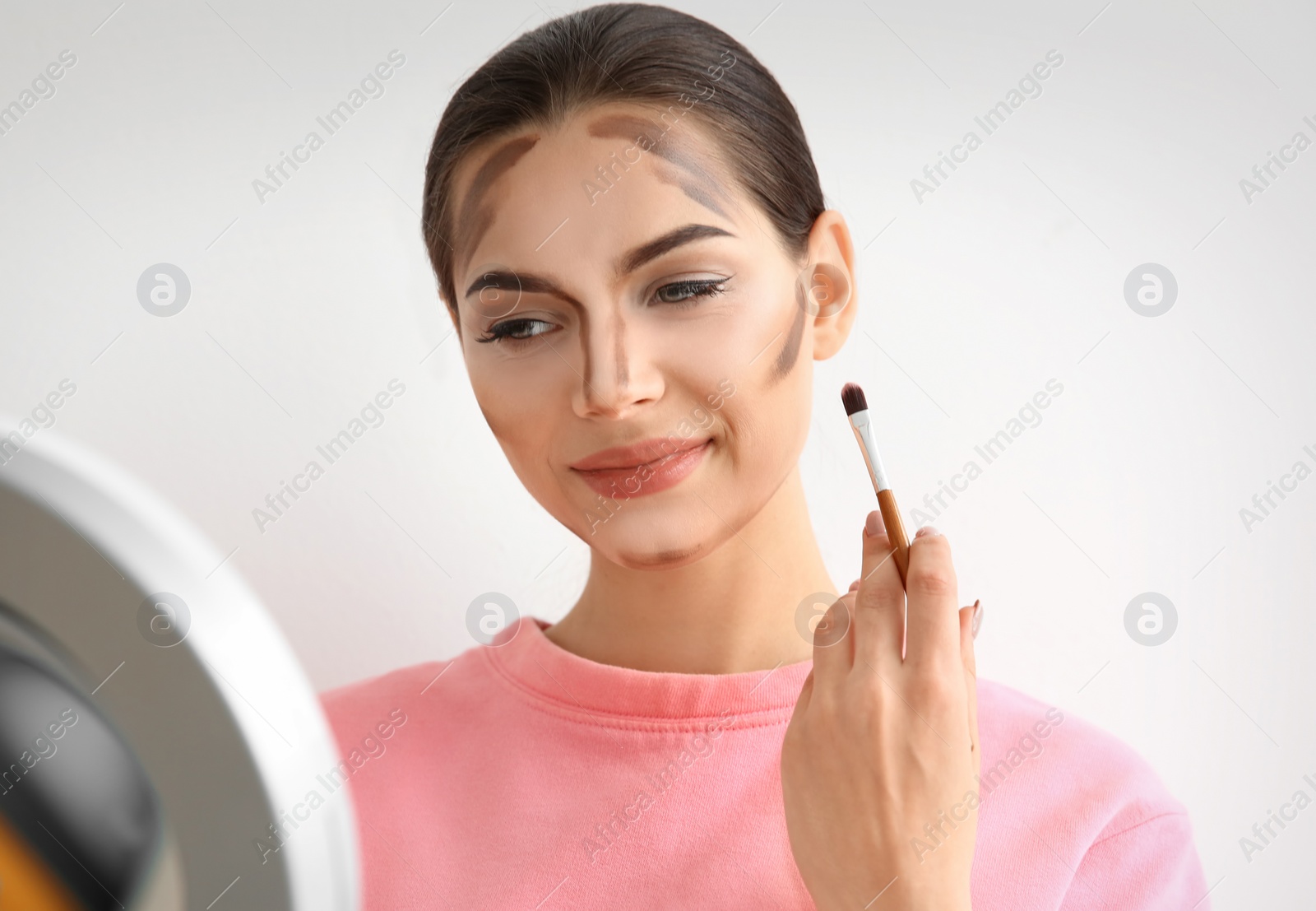 Photo of Young woman applying contouring lines on her face against light background. Professional makeup products