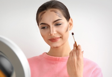 Photo of Young woman applying contouring lines on her face against light background. Professional makeup products