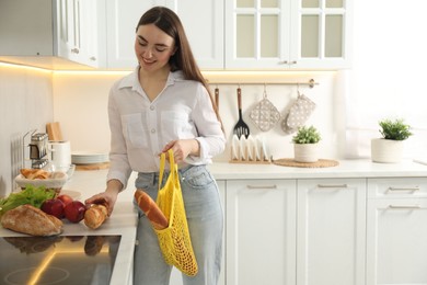 Woman holding string bag with baguette and putting bread on countertop in kitchen