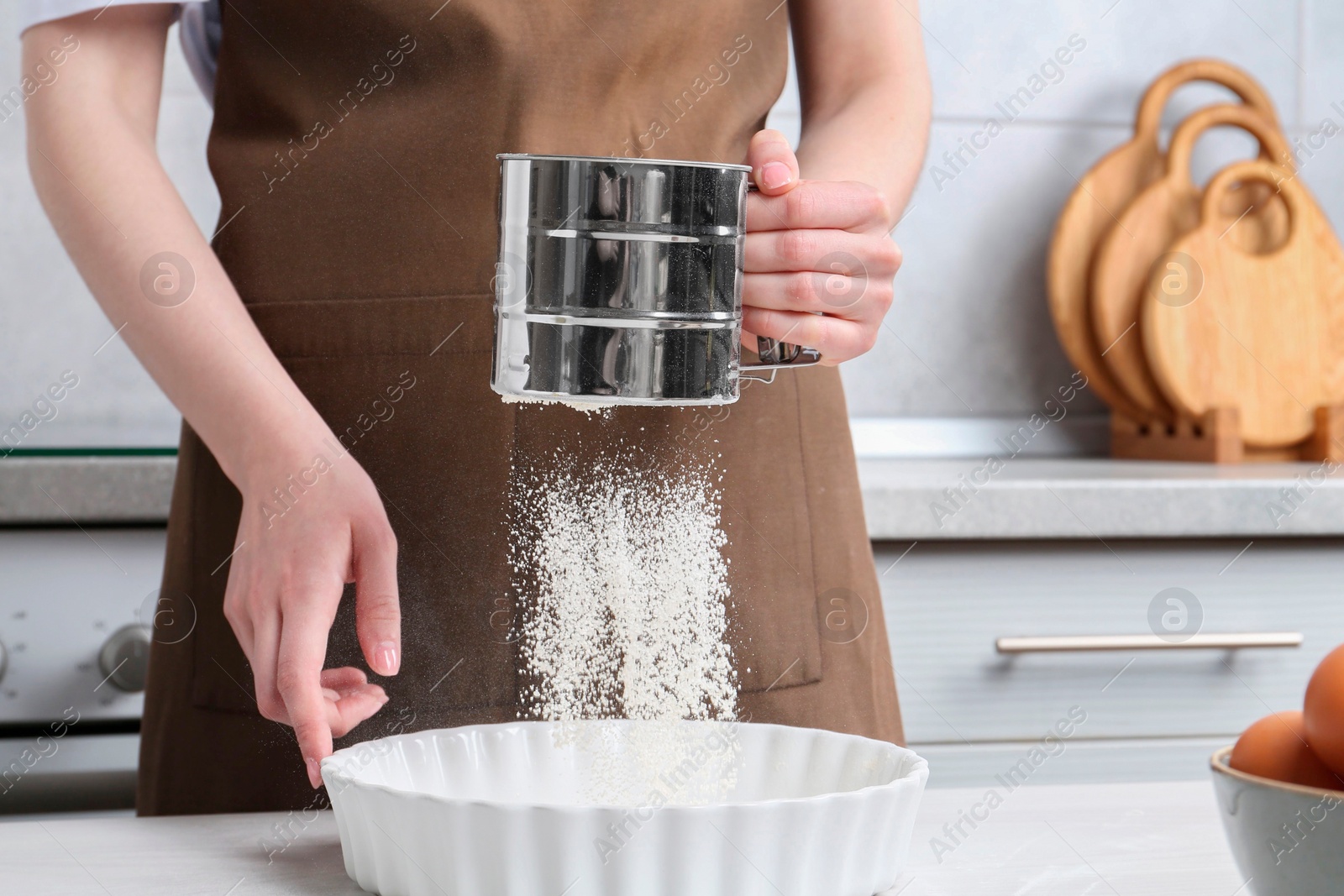 Photo of Woman sieving flour into baking dish at table in kitchen, closeup