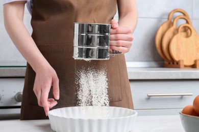 Woman sieving flour into baking dish at table in kitchen, closeup