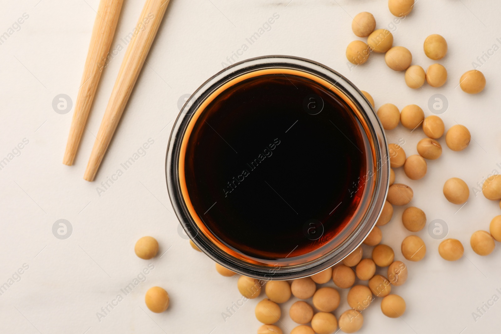 Photo of Soy sauce in bowl, soybeans and chopsticks on white table, flat lay