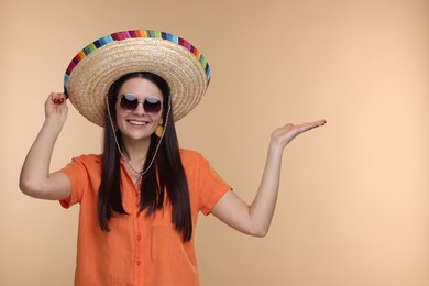 Photo of Young woman in Mexican sombrero hat and sunglasses showing something on beige background. Space for text