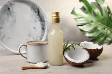 Photo of Bottle of delicious syrup, halves of coconut, flakes, cup of coffee and green leaves on white wooden table